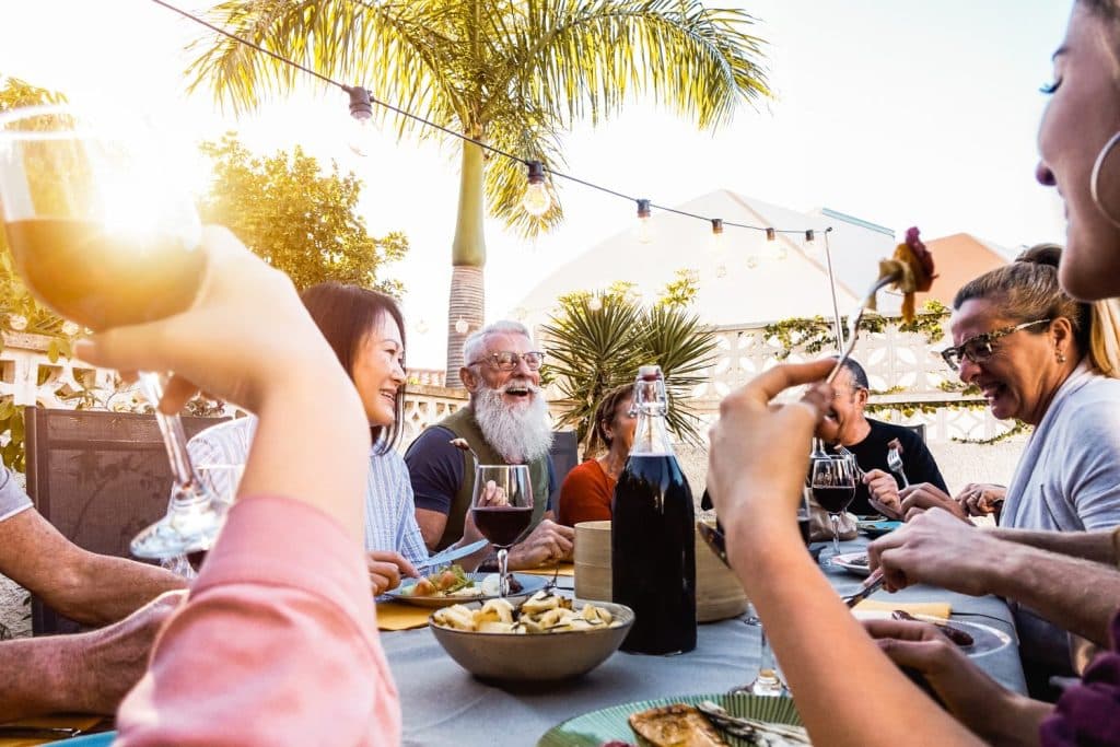 A group of people sit around an outdoor table, eating and drinking, with palm trees and string lights in the background on a sunny day.