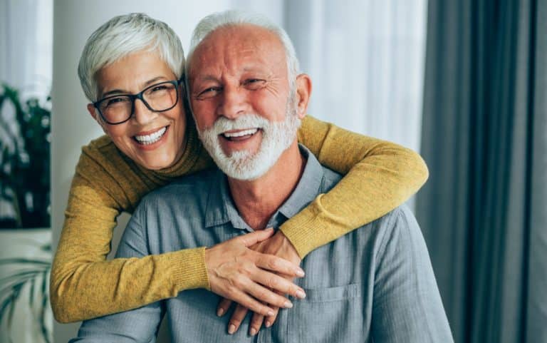 An older woman with short gray hair and glasses hugs a smiling older man with a white beard. Both are indoors and appear happy.