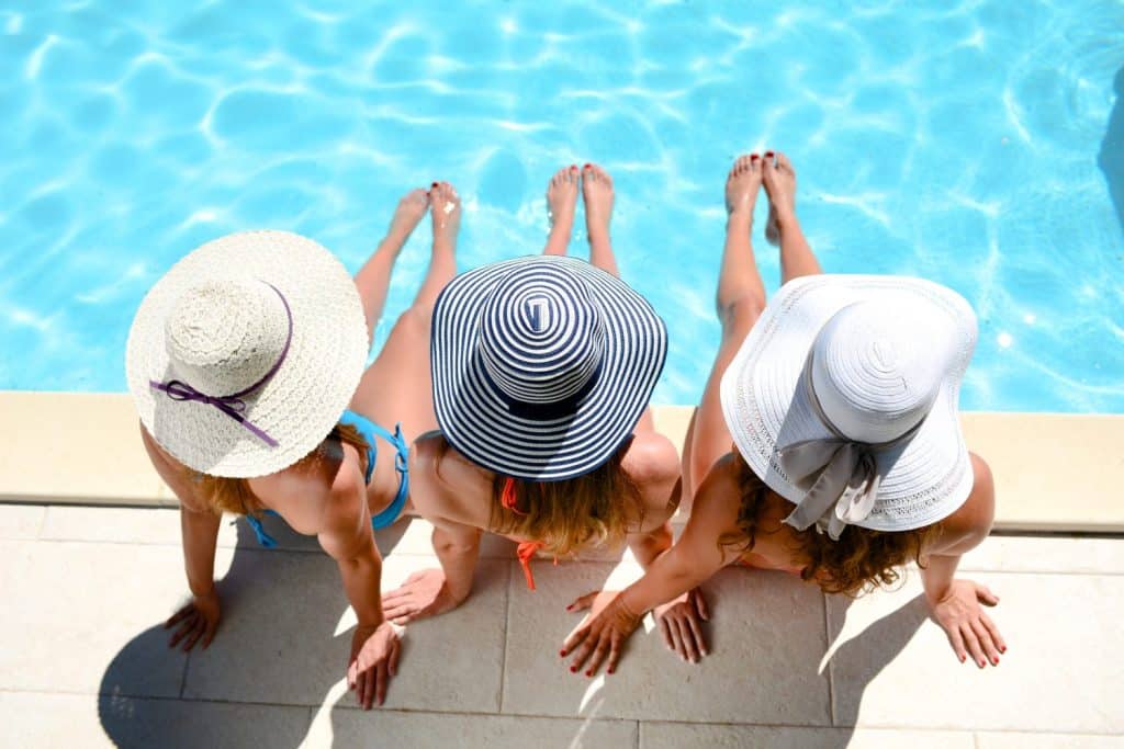 Three women in swimsuits and sun hats sit on the edge of a swimming pool with their legs in the water, viewed from above.