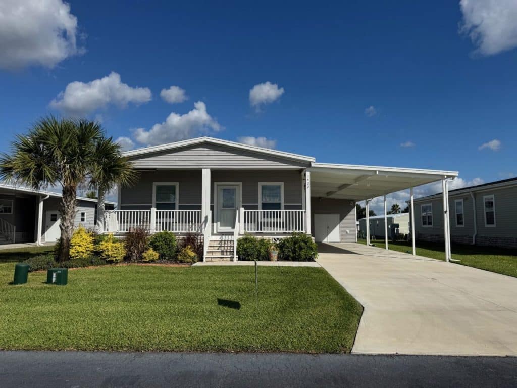 A single-story manufactured home with a front porch, carport, lawn, and palm tree under a blue sky with scattered clouds.