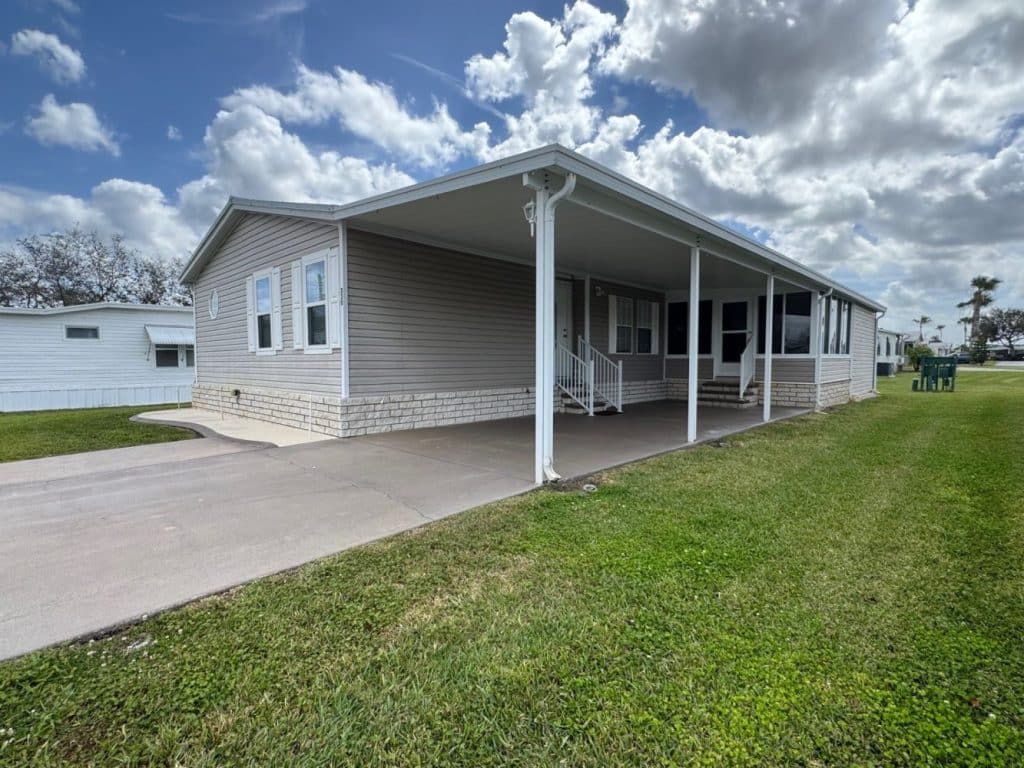 A single-story manufactured home with a covered carport, beige siding, and white trim sits on a well-maintained grassy lawn under a partly cloudy sky.
