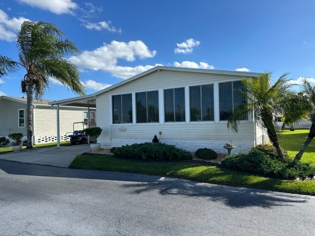 A beige manufactured home with large front windows, a carport, palm trees, and neatly trimmed bushes under a blue sky with scattered clouds.