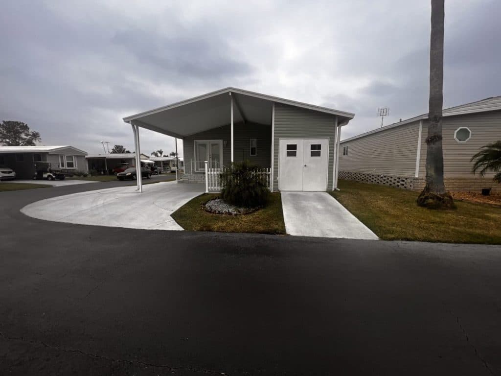A single-story manufactured home with a covered carport, small porch, attached shed, and neatly mowed lawn on a cloudy day.