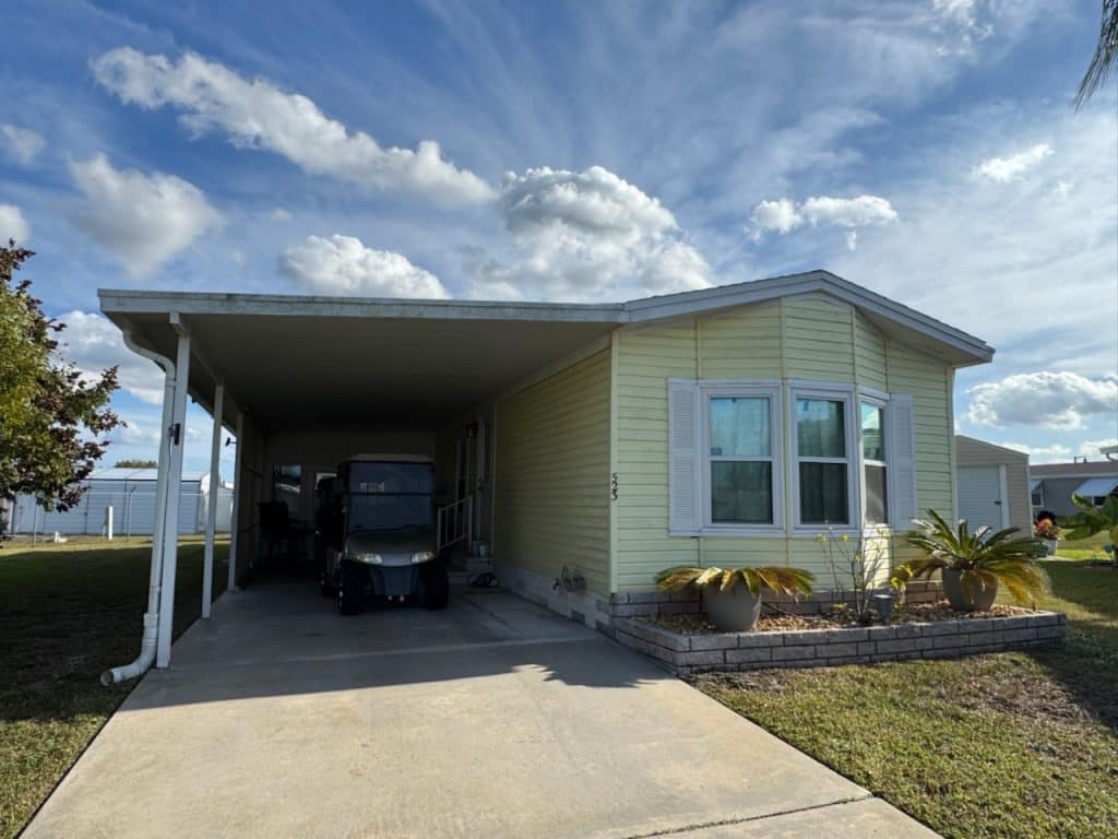 A light yellow manufactured home with white trim, a covered carport, driveway, golf cart, and small landscaped area with potted plants in front.