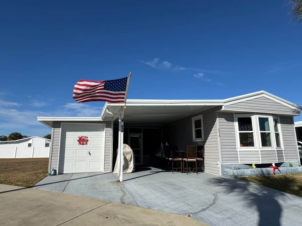 A single-story gray manufactured home with an American flag on a pole, a small porch with chairs, and a driveway under a clear blue sky.