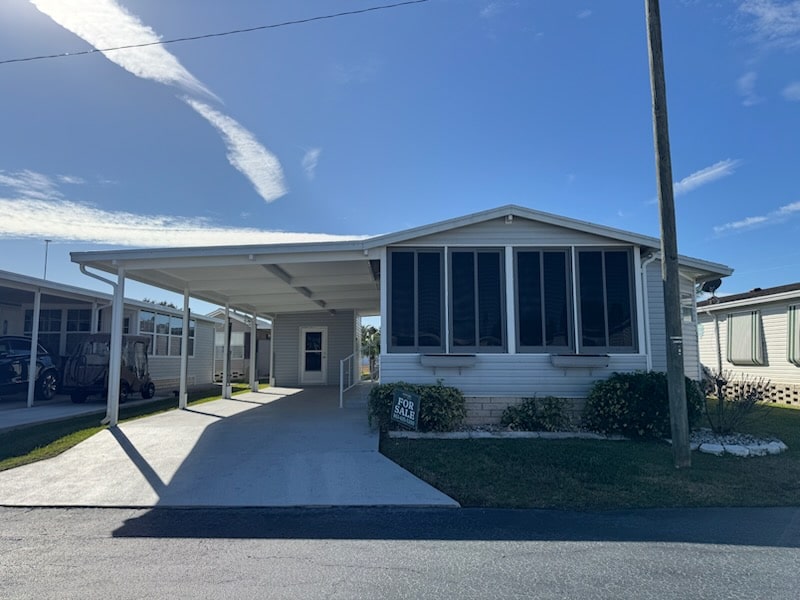 Single-story manufactured home with large windows, a carport, and a "For Sale" sign on the front lawn, set on a sunny day in a residential community.