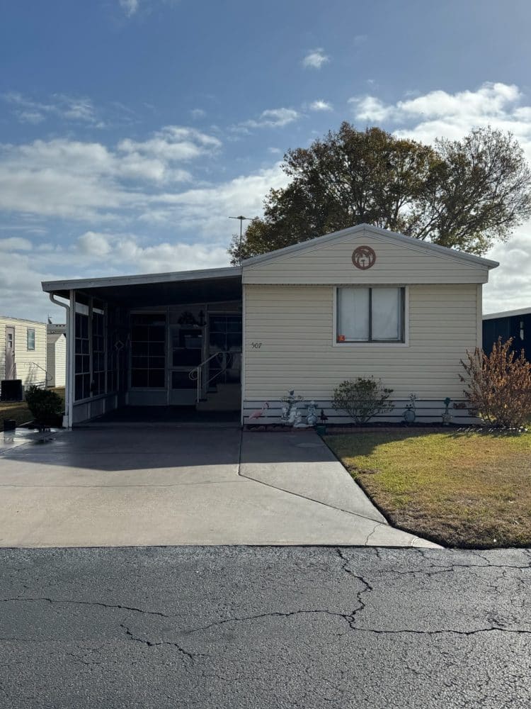 A beige mobile home with an attached covered carport, small front yard, concrete driveway, and a few plants, under a partly cloudy sky.