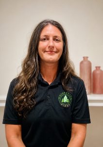 Woman with long brown hair in a black polo shirt, smiling, with two vases in the background.
